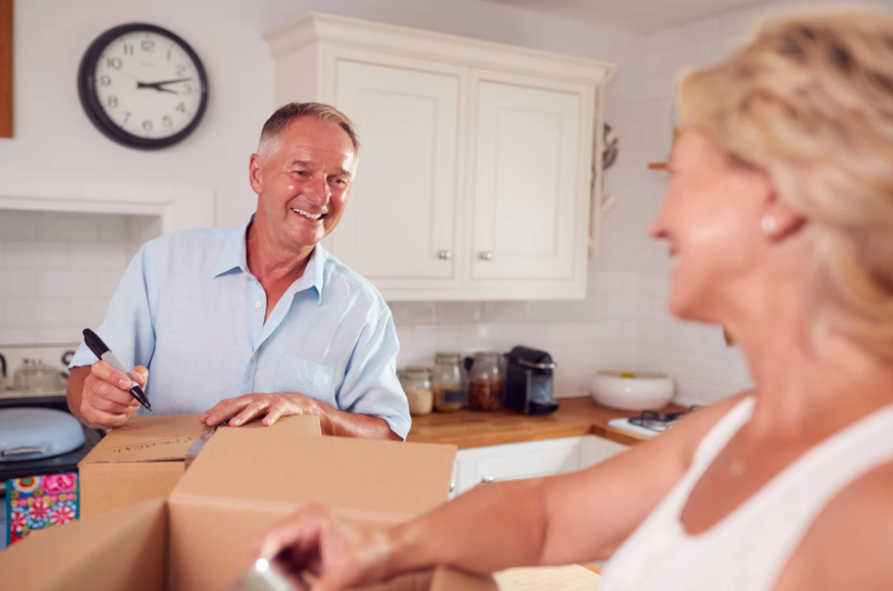 A smiling senior couple packs boxes together in a bright kitchen, capturing a lighthearted moment during their downsizing process.
